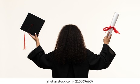 Back View Unrecognizable Woman Wearing Ceremony Robe Holding Certificate And Graduation Cap On Beige Background. Girl Celebrating Graduation And Getting Diploma. Graduate Cap And Degree Paper