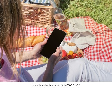 Back View Of Unrecognizable Woman In White Pants Outside Having Picnic And Using Smartphone Taking Photo. Summer Fun And Leisure. View From Behind