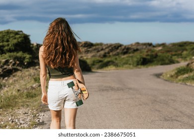 Back view of unrecognizable young female skateboarder with longboard strolling on walkway against mountain and sea - Powered by Shutterstock