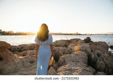 Back view of unrecognizable young woman traveler with long hair in stylish maxi dress with straw hat in hand standing on massive rocky boulders. She is admiring sea against cloudless sunset sky 