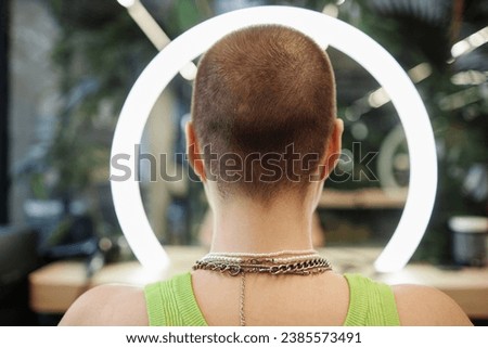 Back view of unrecognizable young woman with buzzcut looking at ring light in beauty salon taking before and after pictures