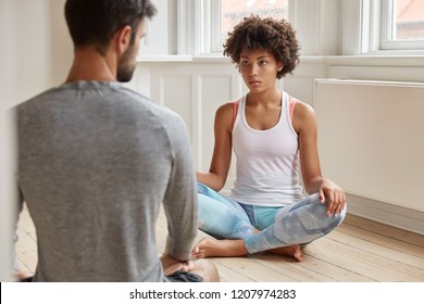 Back view of unshaven man has talk with dark skinned woman, sit on floor together in lotus pose in empty room. Diverse woman and man practice yoga indoor. Ethnicity and spirituality concept. - Powered by Shutterstock