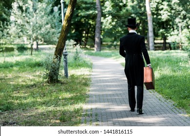back view of victorian man in hat holding baggage and walking outside 