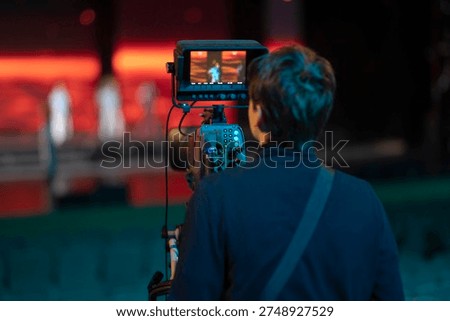 Back view of a videographer operating a broadcasting camera during a stage show with red lighting. Concept of media production, filmmaking, and event coverage