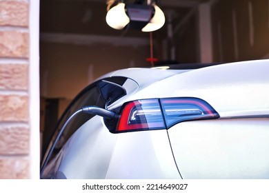 Back View Of White Modern Electric Car Plugged And Charging In Garage At Home Against Blurred Garage Opener And Wall Interior Background. Home EV Charger Station Is Convenient And Cost Efficient. 