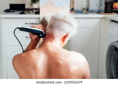 Back view white, silver hair man doing self haircut with a clipper and looks in the mirror. male self-care at home with electric razor. Selective focus. - Powered by Shutterstock