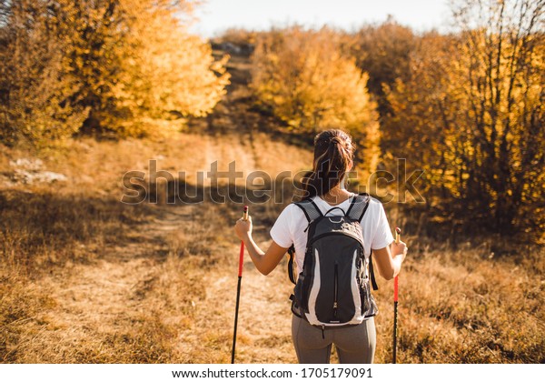 Back view of woman with backpack and trekking\
sticks. Nordic walking. and hiking. Autumn nature around and path\
forward. Travel concept.