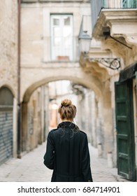 Back view of woman in black classic coat standing on background of old narrow street. 