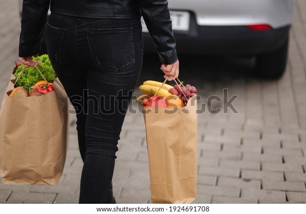 Back view of woman in black clothes carries a\
eco package full of griceries to the car. Fresh vegetables and\
fruits. Midle selection