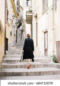 Back View Of Woman In Black Coat And Shoes Walking Up Stairs On Narrow Street. 