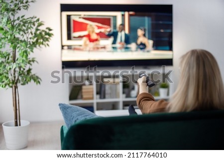 Back view of woman with blond hair sitting on comfy couch and using remote control for wathing TV. Leisure activity at home of caucasian female person.