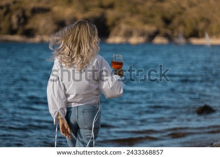 Back view of a woman with blonde hair, holding glass of red wine and looking out over water. A lady wearing white shirt and jeans looking out over blue water with a glass of red wine in the sunshine.
