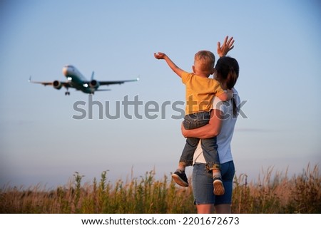 Back view of woman with boy waving hands to landing commercial airplane at sunset. Lifestyle and travel concept.