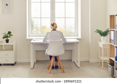 Back View Of Woman Busy With Paperwork, Sitting At Table By The Window In Hospital Or Clinic Office. Doctor Working At Desk, Filling Out Medical History Or Doing Scientific Research On Computer