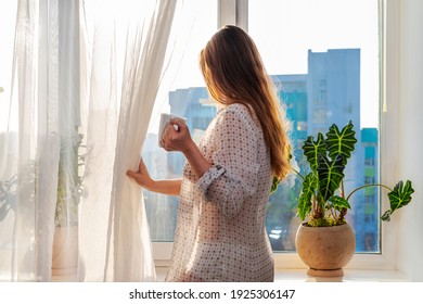 Back View Of A Woman In A Casual Shirt With Long Brown Hair With A Cup Of Tea Or Coffee Looking Out Of The Window, Pulling Back The Curtain. Lifestyle At Home, Natural Sunbeam.