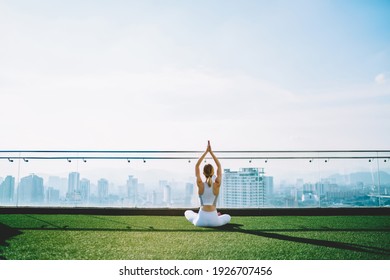 Back view of woman doing yoga Sukhasana with raising hands on lawn on roof of big building in sunny summer morning