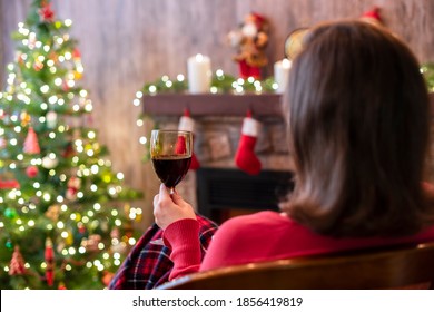 Back View Of Woman Drinking Wine From Glass Covered Plaid Sitting And Relaxation On Armchair Near Christmas Fareplace And Fir-tree. 