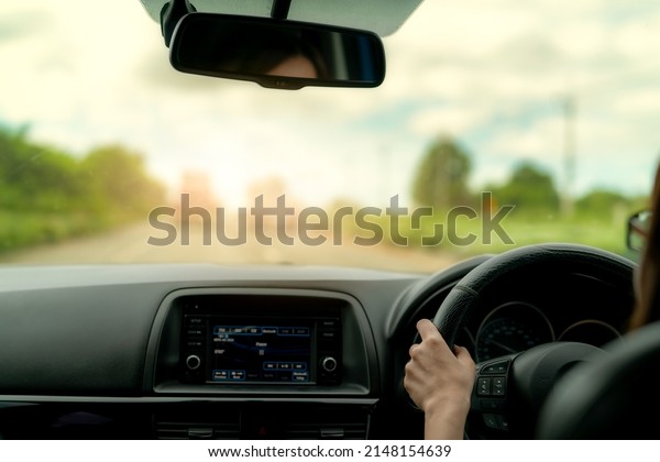 Back view of a woman driving car for summer
road trip travel. Car driving with safety on asphalt road. Driver
hand holding steering wheel for control car. Inside view of car.
Dashboard and windshield.