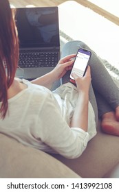 Back View Of A Woman Hand Holding A Smart Phone With Blank White Screen Lying On A Sofa At Home.