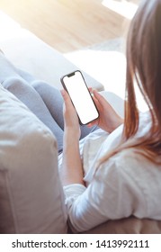 Back View Of A Woman Hand Holding A Smart Phone With Blank White Screen Lying On A Sofa At Home.