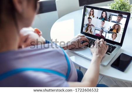 Back view of woman and her baby son doing online meeting with coworkers on video call on laptop at home.