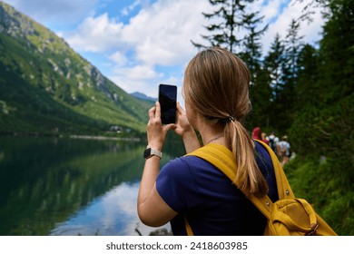 Back view of woman hiking near mountain lake with backpack, taking photo with smartphone. Female traveler walking in forest. Traveling in national park. Active recreation - Powered by Shutterstock