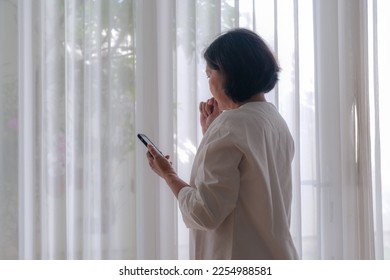 Back view: A woman holding a cell phone standing near the window; biting nails. - Shutterstock ID 2254988581
