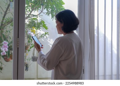 Back view: A woman holding a cell phone standing near the window - Shutterstock ID 2254988983