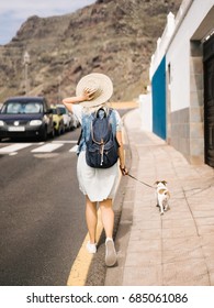 Back view of woman holding hat and walking dog on a town street. 