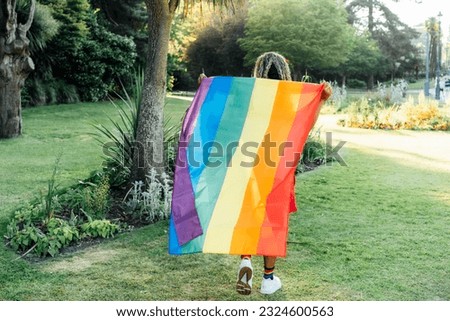 Back view woman holding a rainbow flag outdoor. Young lesbian activist walking with flag symbol of social movement Lgbtq. Fight for equality, freedom and human rights. Celebrating Pride month.