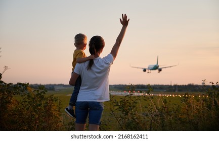 Back View Of Woman With Kid Waving Hands To Landing Commercial Airplane At Sunset. Lifestyle And Travel Concept.