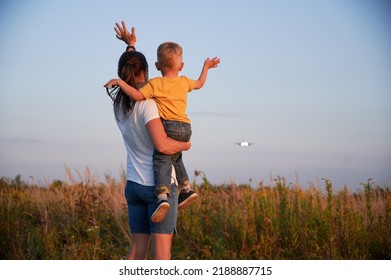 Back View Of Woman With Kid Waving Hands To Flying Commercial Airplane In The Sky At Sunset. Lifestyle And Travel Concept.