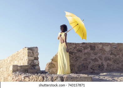 Back View Of Woman In Long Fashion Dress Standing On Stone Architecture And Holding Yellow Umbrella. Bad And Wrong Photoshop Edit.