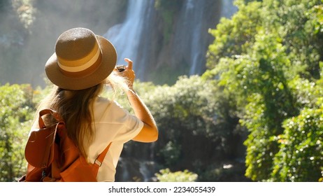 Back view of woman with photo camera shooting tropical waterfall. Inspiring millennial travel blogger in journey. Happy young traveler girl on summer vacation. Concept of wanderlust and adventure - Powered by Shutterstock