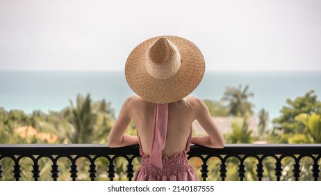 Back View Of Woman In Pink Dress And Straw Hat Standing On A Hotel Balcony, Ocean View.