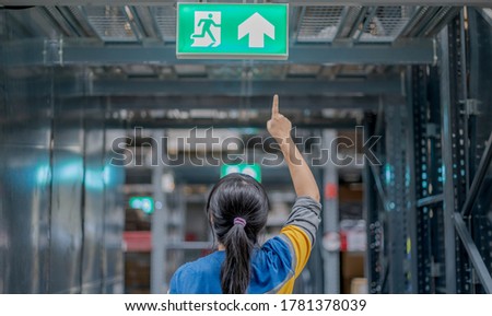 The back view woman pointing at the emergency fire exit sign show the way to escape at the public area, warehouse store. For the security first concept.