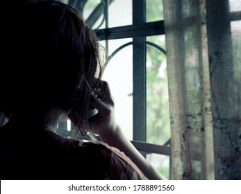 Back view of a woman receiving a foreboding call. Silhouette of lady by the window at home. Distress, loss, or sadness concept.