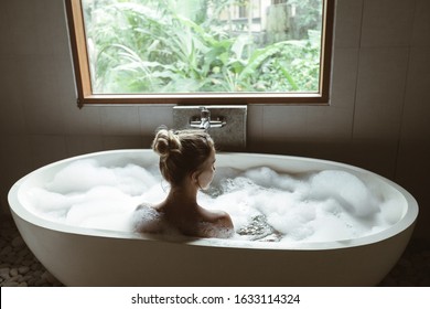 Back View Of Woman Relaxing With Pleasue In Foam Bath With Bubbles In Modern Hotel Dark Bathroom By Big Window