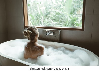 Back View Of Woman Relaxing With Pleasue In Foam Bath With Bubbles In Modern Hotel Dark Bathroom By Big Window