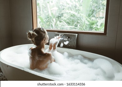 Back view of woman relaxing with pleasue in foam bath with bubbles in modern hotel dark bathroom by big window - Powered by Shutterstock