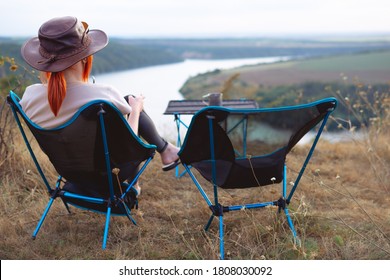 Back View. A Woman Sits On A Folding, Portable Chair And Enjoys The Moment Of Life. Nature, Mountains And River, Sunset. Tourism And Active Lifestyle. Equipment For Tourism.