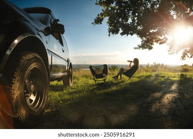 Back view. A woman sits on a folding, portable chair and enjoys the moment. Nature, on the top, sunset rays. Tourism and active lifestyle. Equipment for tourism. Off road vehicle wheels - Powered by Shutterstock