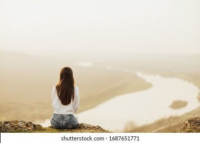 Back view of  woman sitting on top of the hill and looking at river in evening sunset. Young  traveler enjoying time in nature. Lifestyle travel, relaxation and freedom concept. Copy space. 