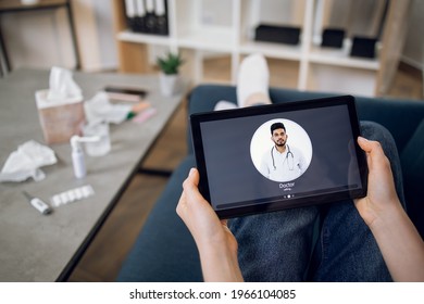 Back View Of Woman Sitting On Sofa With Digital Tablet, Staying At Home, Making Video Call To Her Male Indian General Practitioner Doctor Via Healthcare App. Sick Girl At Online Consultation