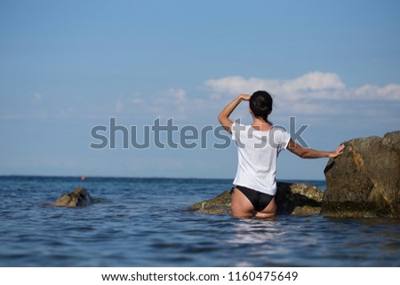 Back view of woman sitting in water next to big rocks looking far away at the sea. Female admiring the blue sea landscape. Holiday vacation concept