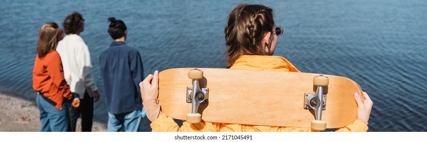 Back View Of Woman With Skateboard Near Blurred Friends And River, Banner