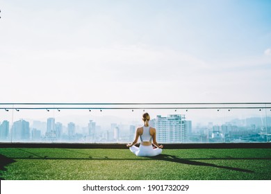 Back view of woman in sports outfit practicing yoga taking Sukhasana pose on top of big building in sunny summer morning