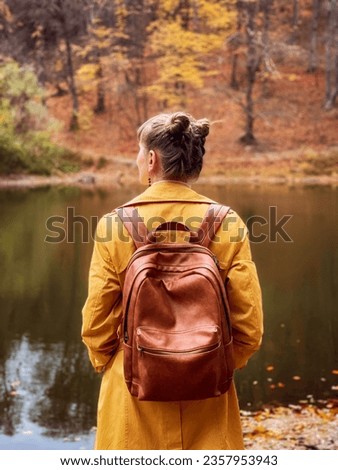 Back view of woman standing in autumn forest and looking at lake. Young female wearing orange coat standing backwards and watching nature. Girl with brown backpack enjoying fall nature colors in park.