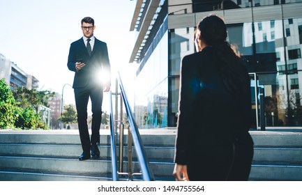 Back View Of Woman In Suit Walking On Stairs With Modern Businessman Using Mobile Phone Walking Opposite In Bright Back Lit