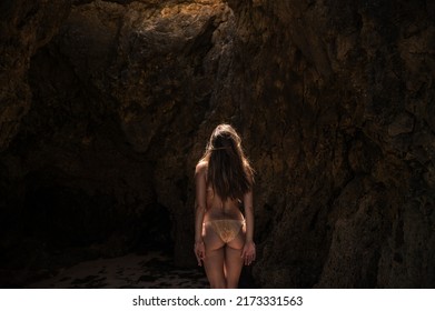 Back View Of Woman In Swimwear Standing On Sandy Coast Near Rocky Formation And Looking Up While Enjoying Sunny Day In Algarve